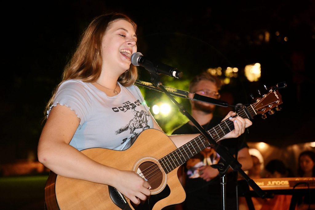 Sarah Gross singing and holding her acoustic guitar a the One with a Concert on the Orange Grove concert