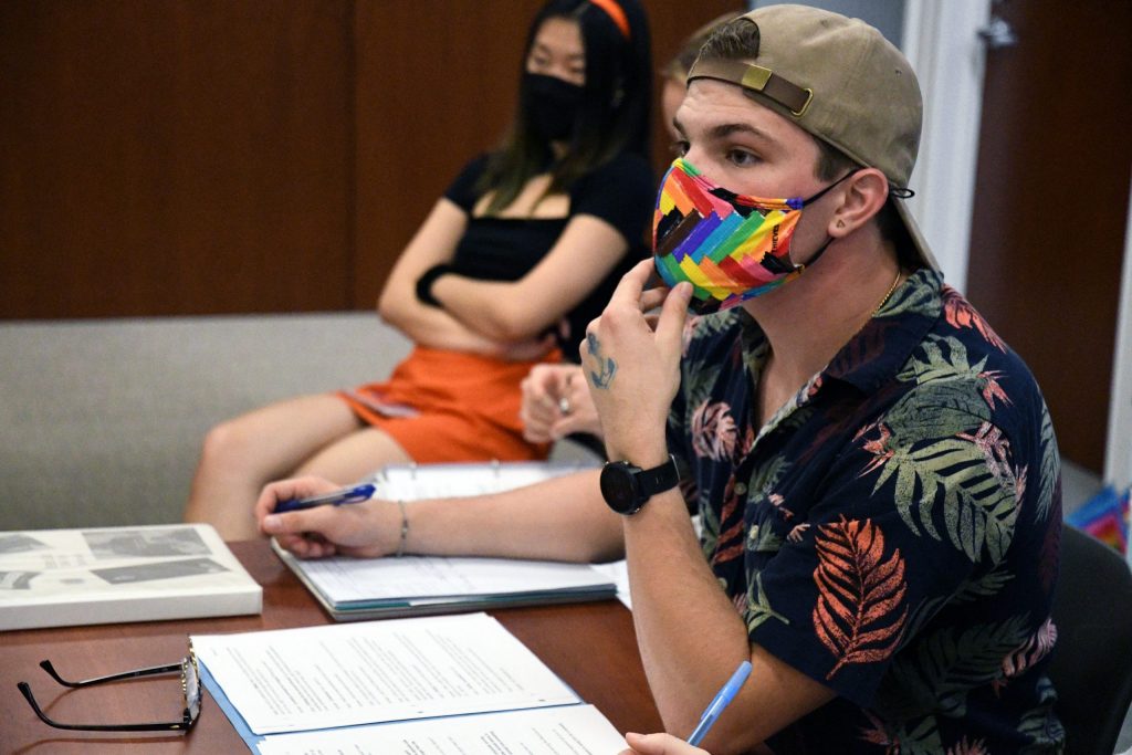 Director Seth Knievel, wearing a backwards hat and rainbow mask, sits at a desk with scripts and notebooks.