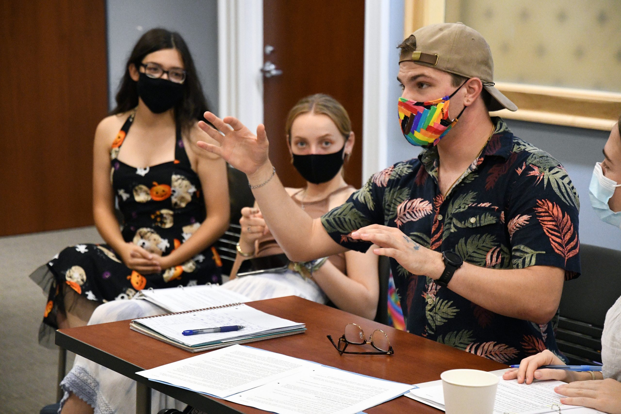 Director Seth Knievel, wearing a backwards hat and rainbow mask, sits at a desk with scripts and notebooks. SOme of the cast sits in the background.