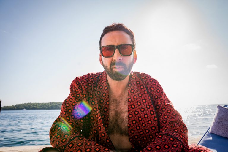 Nicolas Cage sits poolside in a red robe and sunglasses staring at the camera