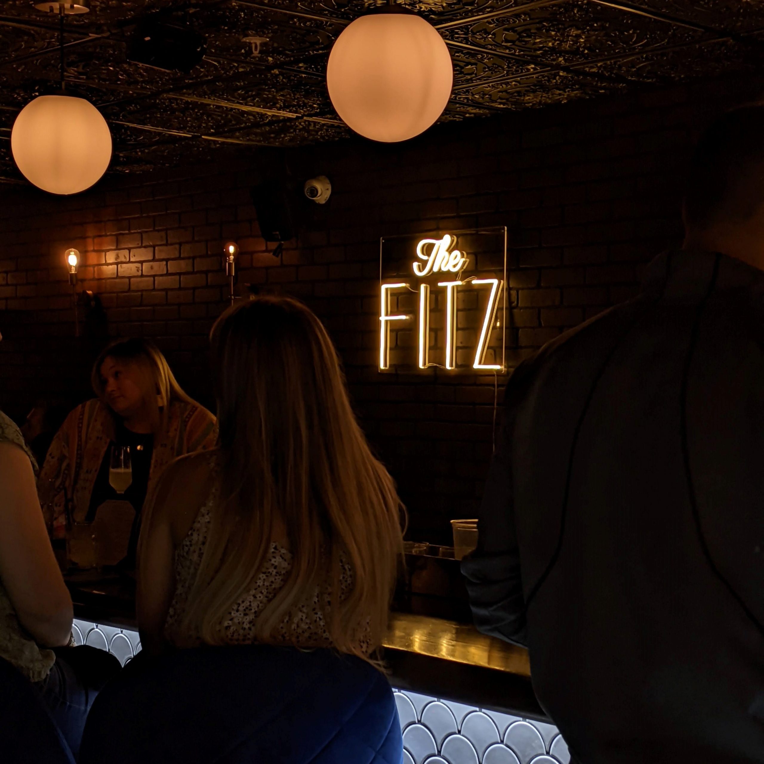 People gather around the bar of the modern speakeasy The Fitz lit by round lamps and a neon sign with the bar's logo.