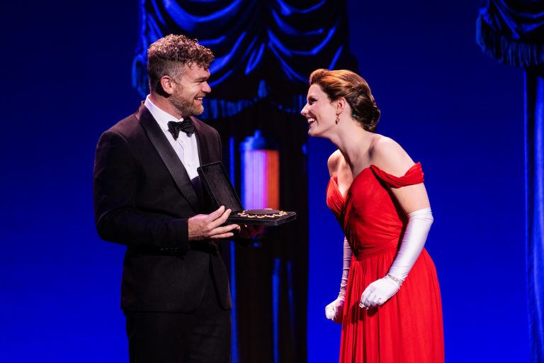 Man in black tuxedo hands a necklace to woman in red dress.
