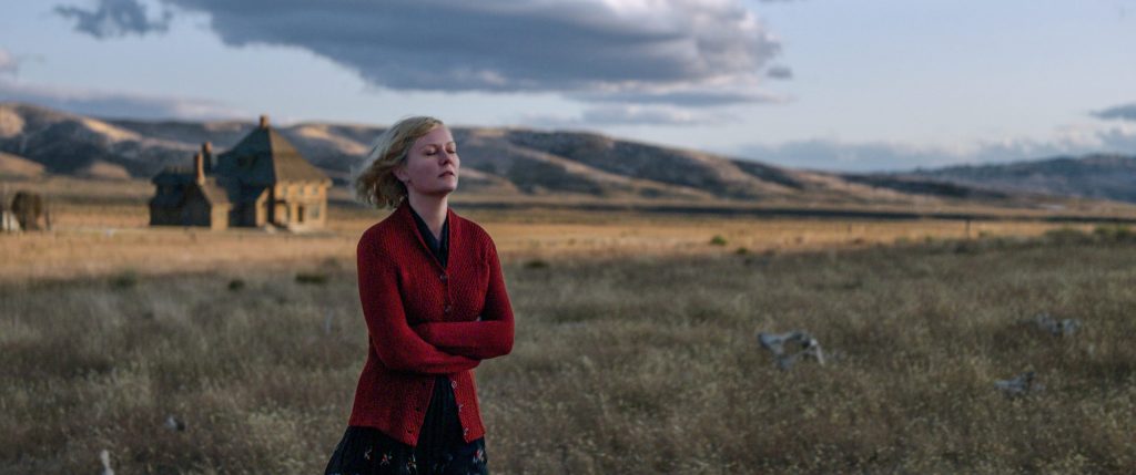 KIRSTEN DUNST stands in a red sweater in a windswept field in film The Power of the Dog