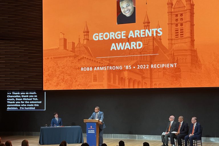 Syracuse Class of '85 alum Robb Armstrong stands at a podium in front of a darkened auditorium, with an orange screen behind him that says "George Arents Award" as well as "Robb Armstrong '85, 2022 recipient".