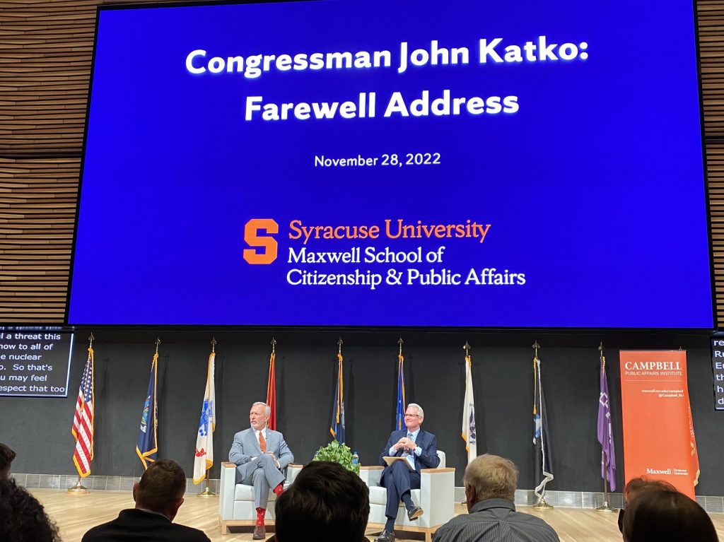 John Katko sits across from Grant Reeher on a stage, both of them facing off stage. A large blue screen behind them reads, 