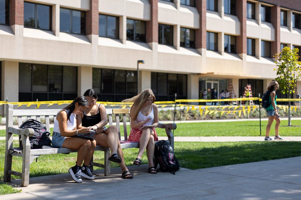 (From left to right) Bridget Sides, Mia Paynton, and Aidan McCarthy sit on a bench together in front of Link Hall on the Quad on .
