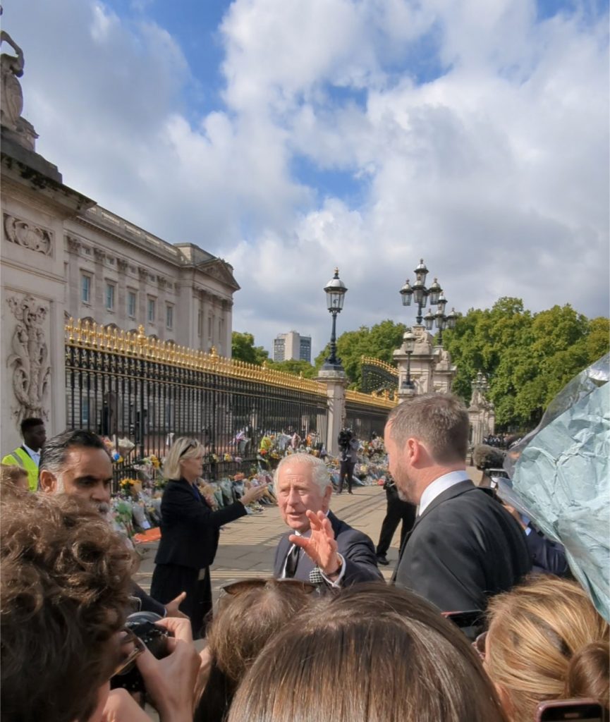 King Charles III, flanked by security, greets people outside of Buckingham Palace, where flowers are linked up against the fence.