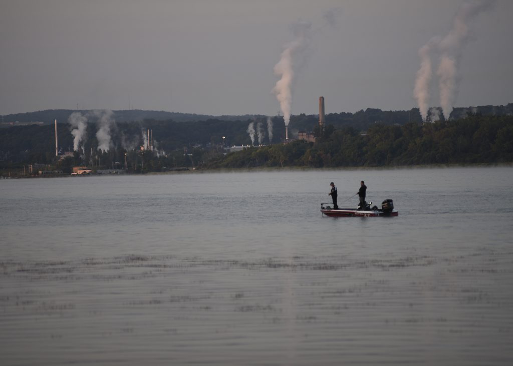 Photo of Onondaga Lake with two people fishing off a boat, and buildings excreting white clouds into the air in the background