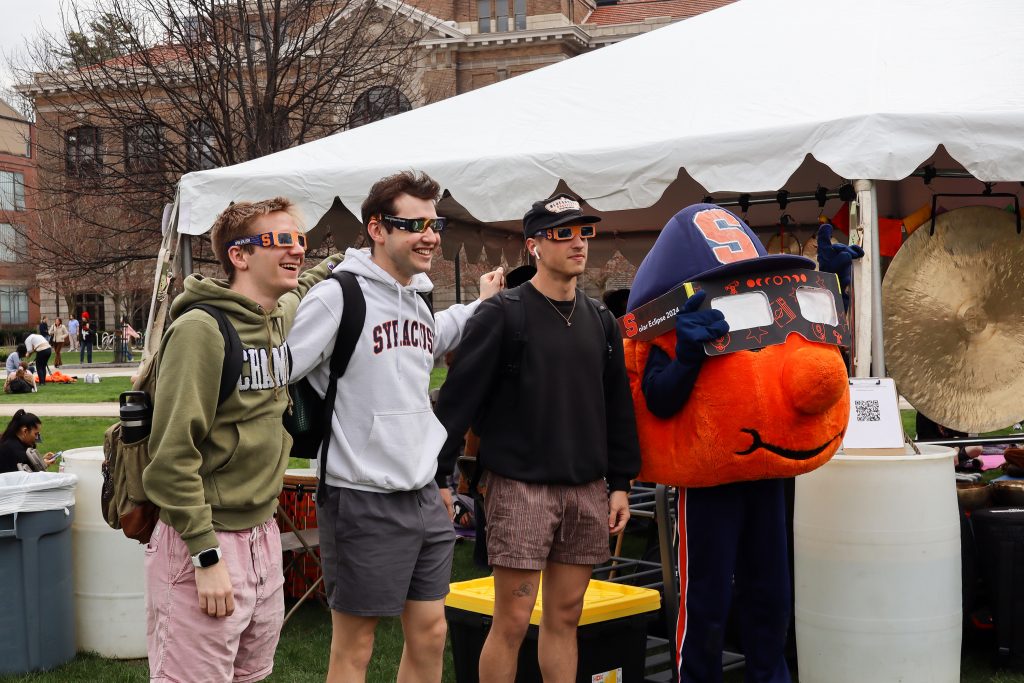 Otto the Orange, Syracuse University's mascot, stands with students wearing eclipse glasses and watching for the rare total eclipse.