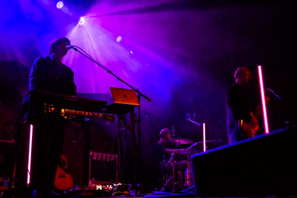 Lights flare through fog as the band Blue October plays. There is a keyboard primarily in frame with the musician playing it obscured in shadow.