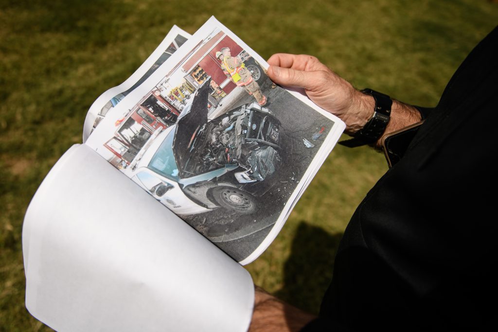 A person holds printed photographs in their hand. The one being shown depicts a police car damaged heavily with the hood up. A firetruck and firemen can be seen in the background of the photo being held.