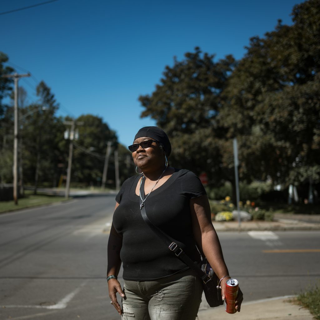 A Black woman stands on the sidewalk at an intersection. She is wearing sunglasses, a black T-shirt, and silver hoop earrings. She is wearing a crossbody bag and holding a canned soft drink.
