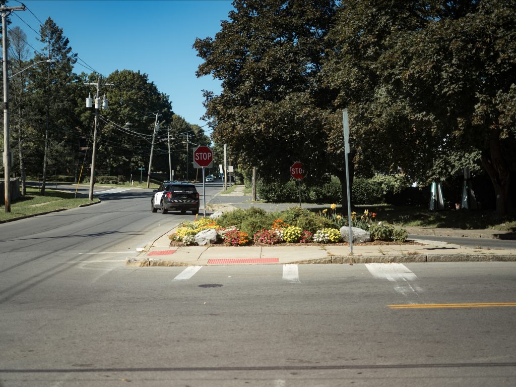 A police SUV turns right onto a two lane road. The camera's view is from an intersection further back. There is an area of flowers as well as two stop signs between the camera and police vehicle.