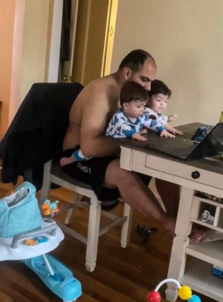 A man sits with his two young sons at a table. The man is holding his two sons in his lap, they are both reaching out and have their hands placed on the table. A laptop and Iphone are placed on the table in front of them.