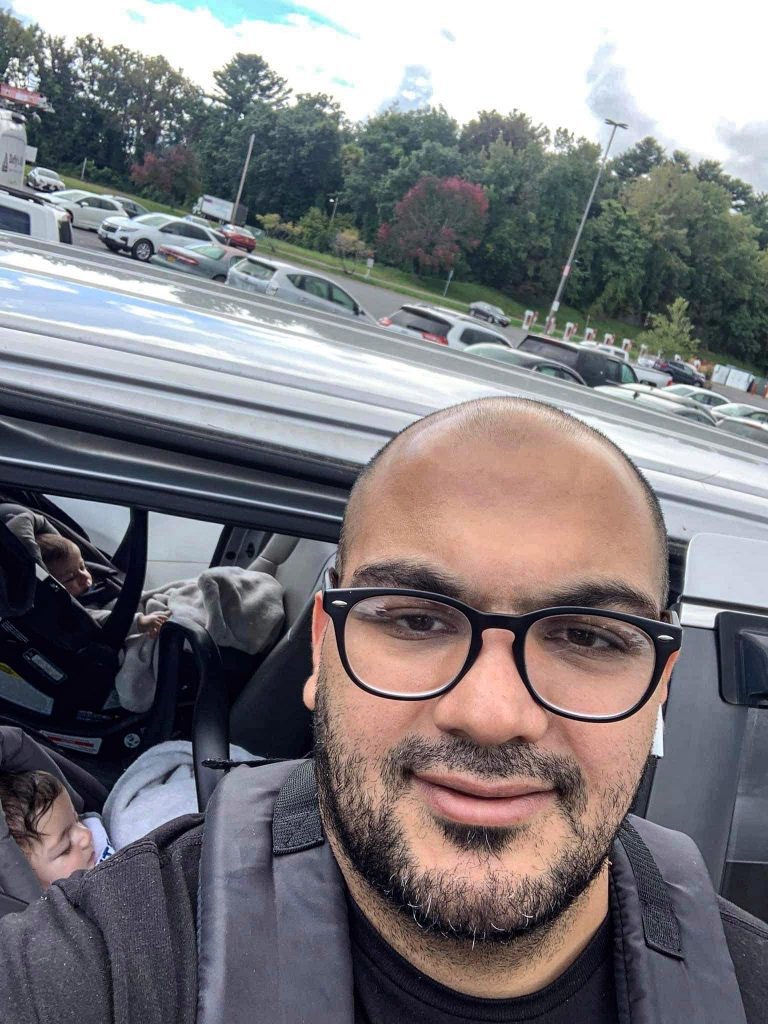 A Selfie of Sabeeh Alikawi and his two infant children who are sitting in his car. Sabeeh is wearing glasses and his twin sons are asleep in the car behind him.