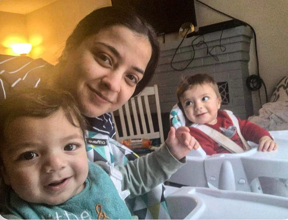 A selfie of a mom and two young twin boys. The boys are sitting in high chairs, one is looking at the camera while the other is looking at his mother. All three are smiling