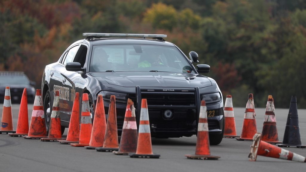 A police car with two passengers drives forward between parallel lines of orange traffic cones. One cone is knocked over ahead of the oncoming vehicle.