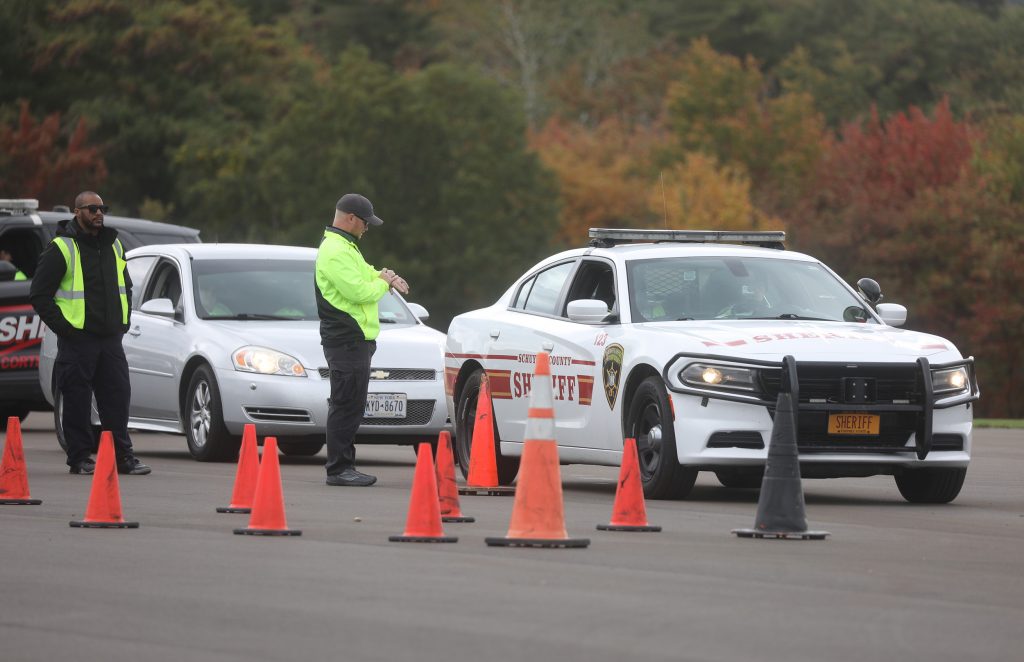 Two men in neon yellow reflective outerwear stand next to a line of three cars, two of which are visibly identifiable as sheriff's vehicles. In front of the men is an arrangement of orange traffic cones. One man appears to be checking his watch.