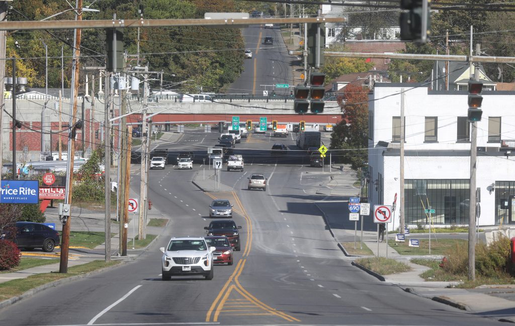 Taken from a high angle in the middle of a four lane, two-way street, cars drive towards and away from the camera. Multiple intersections with stoplights are seen ahead as well as an overpass. A red sign reads, 