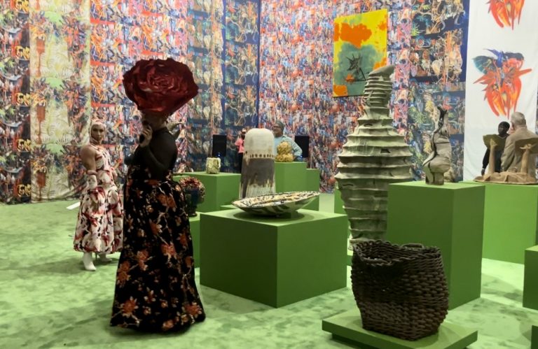 Drag queens Yuka Liptis and Samantha Vega stand to the left of Mexican artist Pepe Mar's "Clay Garden" where green carpeting and pedestal highlight the clay pieces that stand on them. Printed textiles line the walls of the exhibit.