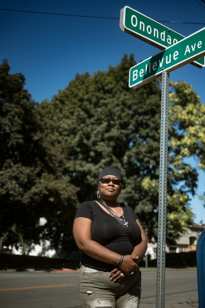 Portrait of a Black woman with the background blurred. She is wearing sunglasses, a black T-shirt, silver hoop earrings, and a silver chain with an angel pendant on it. She is standing next to a street sign that reads 