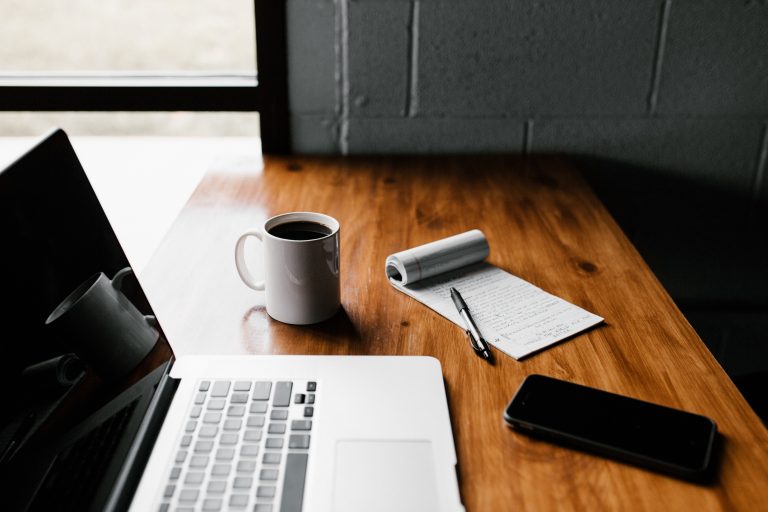 A laptop, coffee mug, notepad and smartphone sitting on a desk.