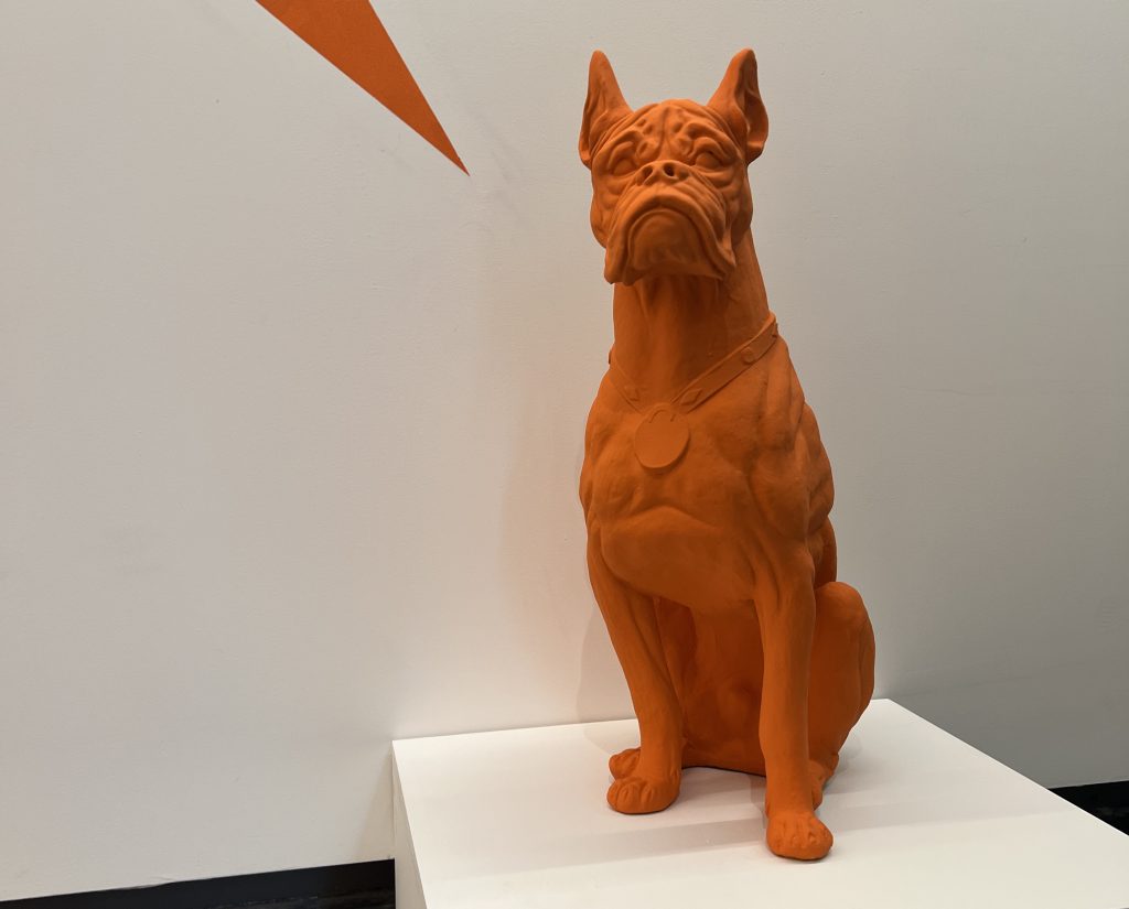 The orange bulldog sits on a white display as a piece of the orange wall behind seemingly points at it.