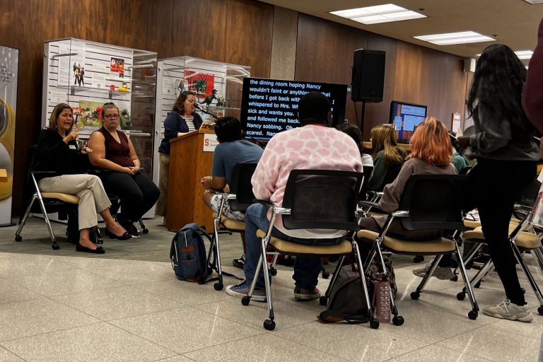 People sitting down on chairs, facing a woman on a podium reading an excerpt from a book, while her words appear on a television screen and a woman sitting near here translates her words to American Sign Language.